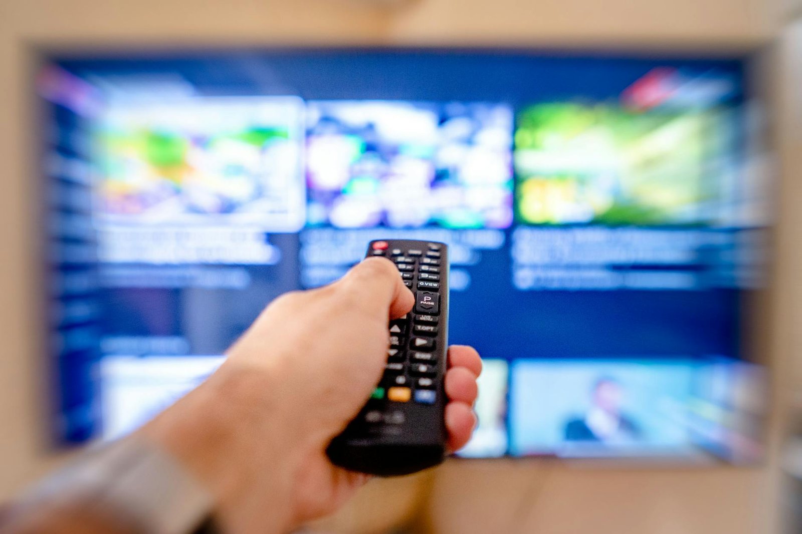 Best IPTV Channels — Hand holding a TV remote pointed at a blurred TV screen in a cozy room | OTT-TV