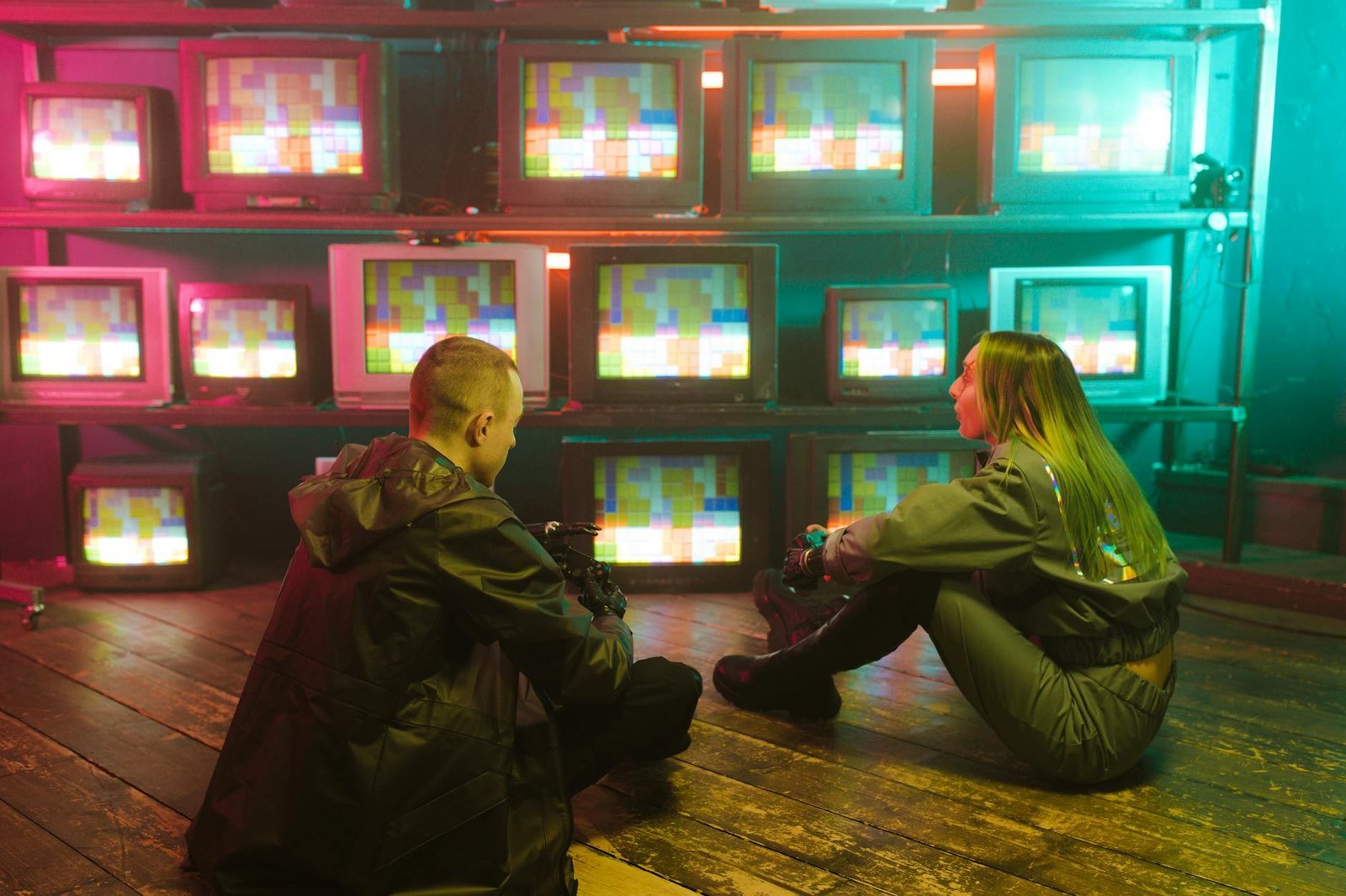 Two people sitting on the floor in front of a wall of vintage CRT televisions glowing with rainbow color bars — most IPTV review sites are paid placements, sort signal from noise