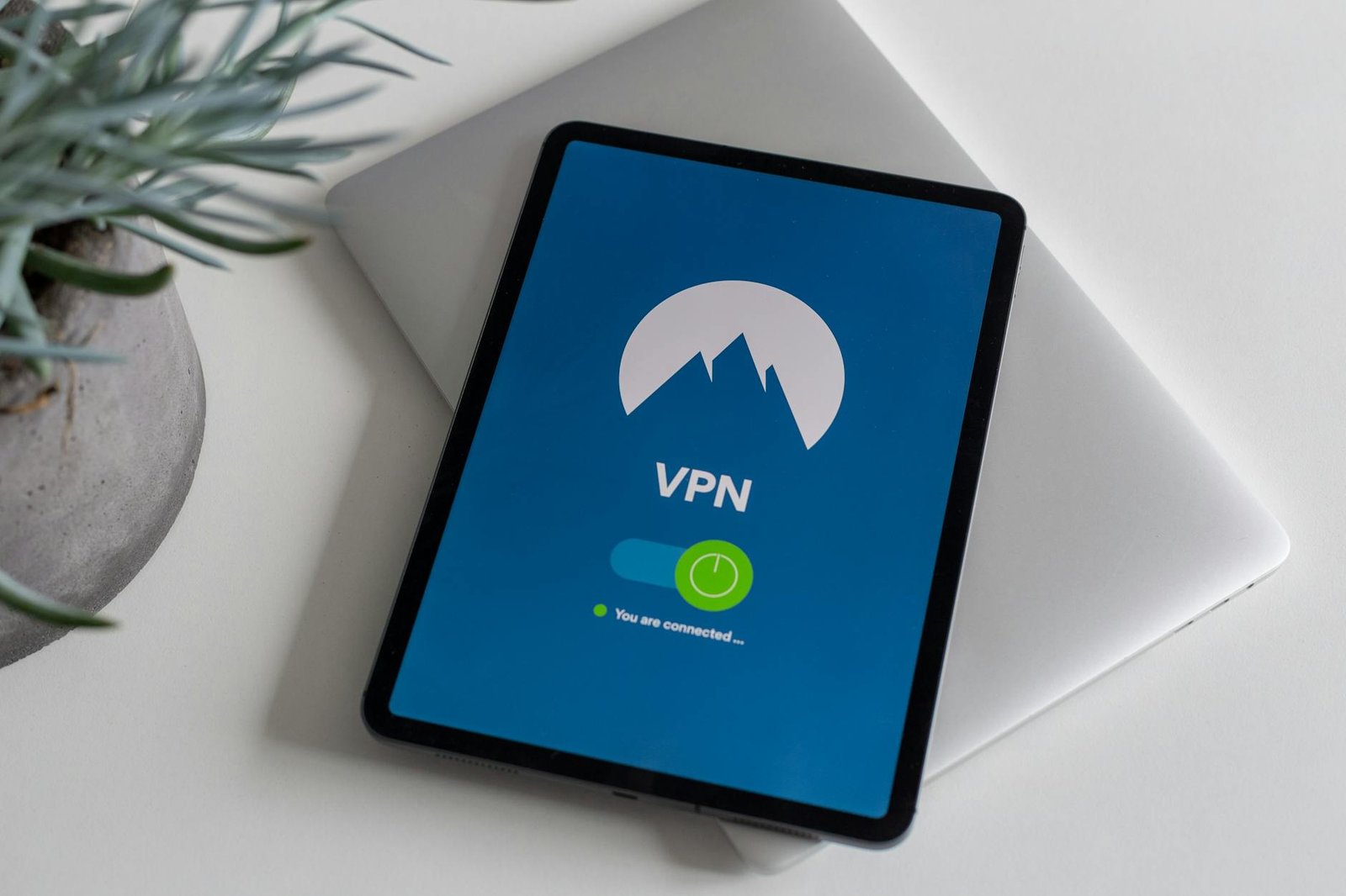 Tablet on top of a closed laptop showing a VPN app screen with a mountain logo and a green "you are connected" indicator — a fast VPN protects IPTV streaming traffic without dropping below 4K bitrates